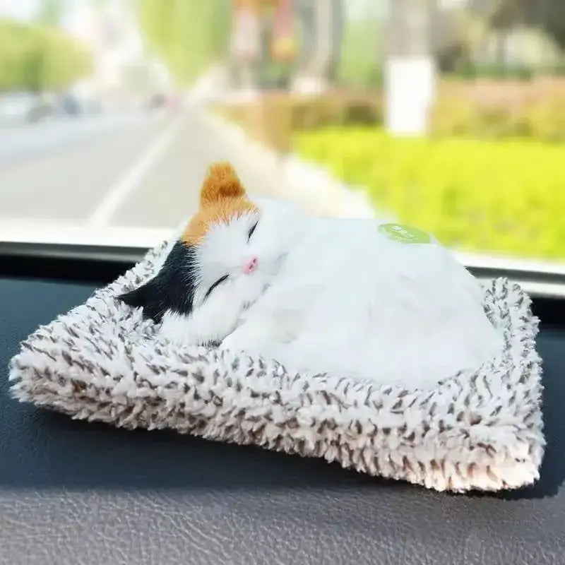 A plush calico cat figurine with white black and orange fur nestled in a soft textured gray and white fuzzy bed as a simulated sleeping cat plush toy with sound.