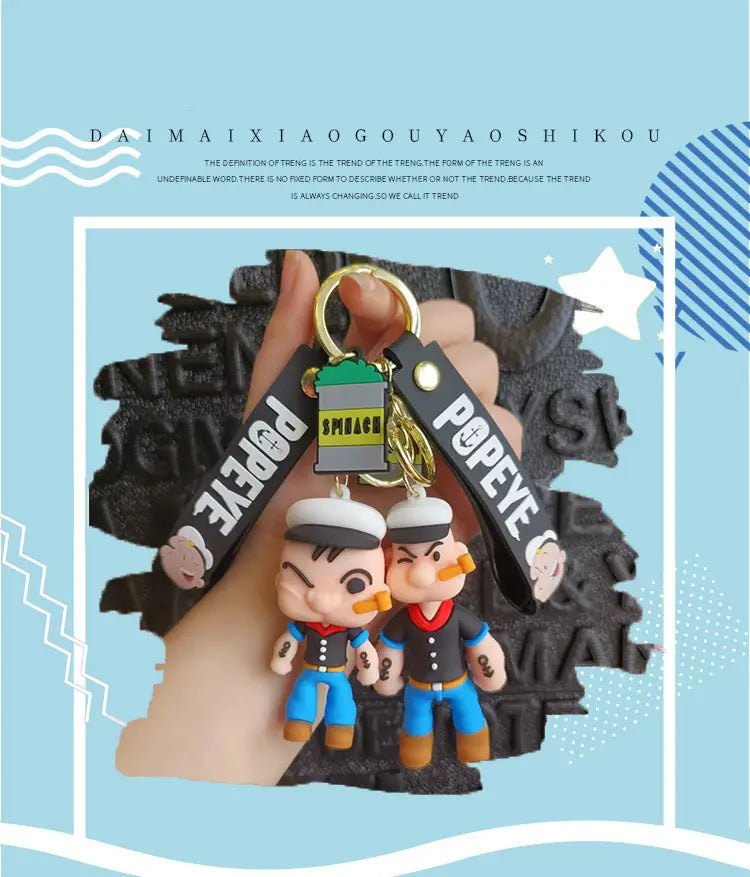 Popeye silicone keychain featuring two cartoonish sailor figures in blue pants and black shirts with white hats attached to a gold-colored ring and black straps bearing the popeye logo.