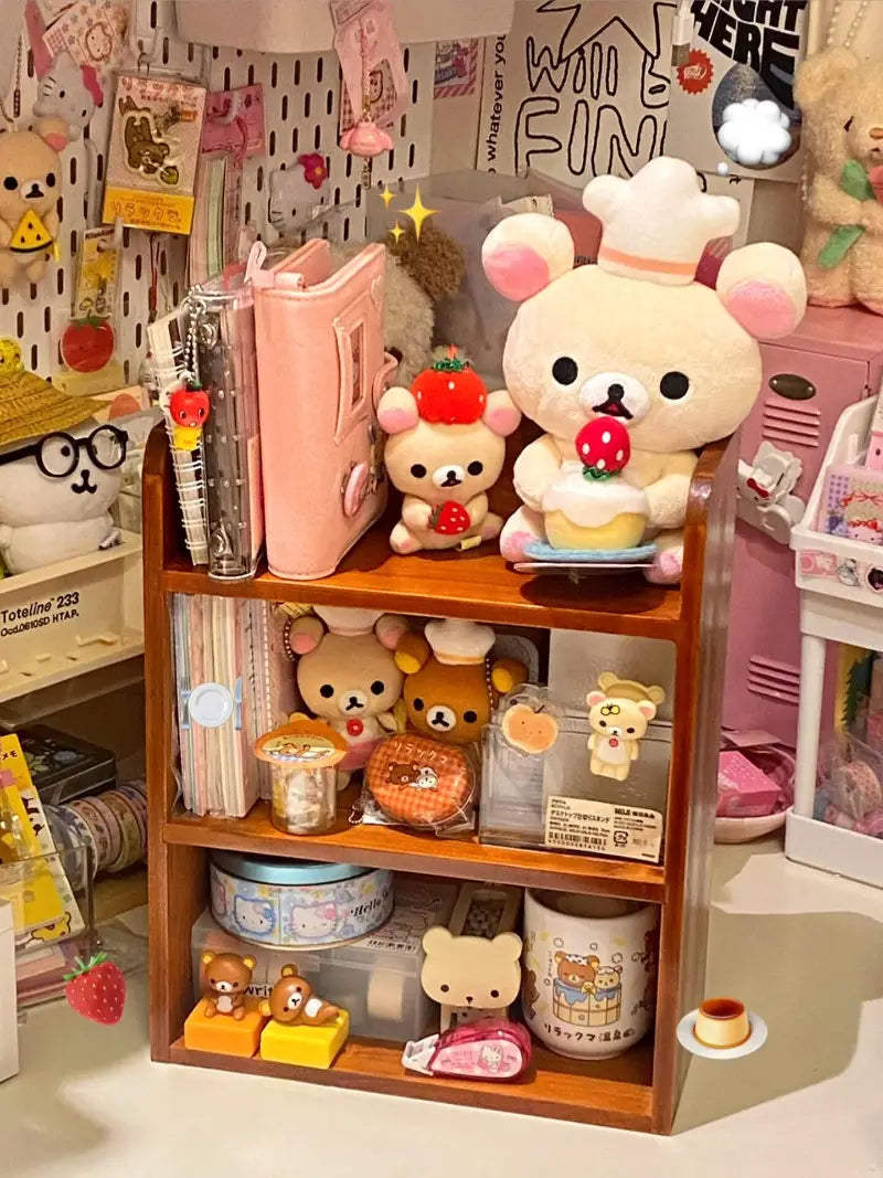 Un personaje rilakkuma esponjoso que lleva un gorro de chef y sostiene un pastel con fresas, sentado sobre una estantería organizadora y para guardar en un escritorio de madera.