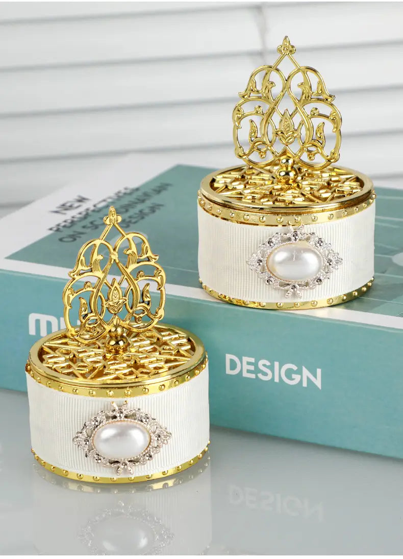 Boîte à bonbons de mariage dorée décorative contenant une perle-bonbon, présentant des boîtes à bijoux ornées de filigrane doré avec des bandes de ruban blanc cannelé, ainsi que la perle centrale et des ornements diamantés.