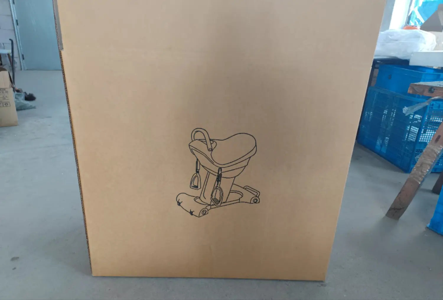 A black line drawing on a brown cardboard box illustrates a horse riding exercise machine fitness equipment featuring handlebars, pedals, and wheels for indoor workout.