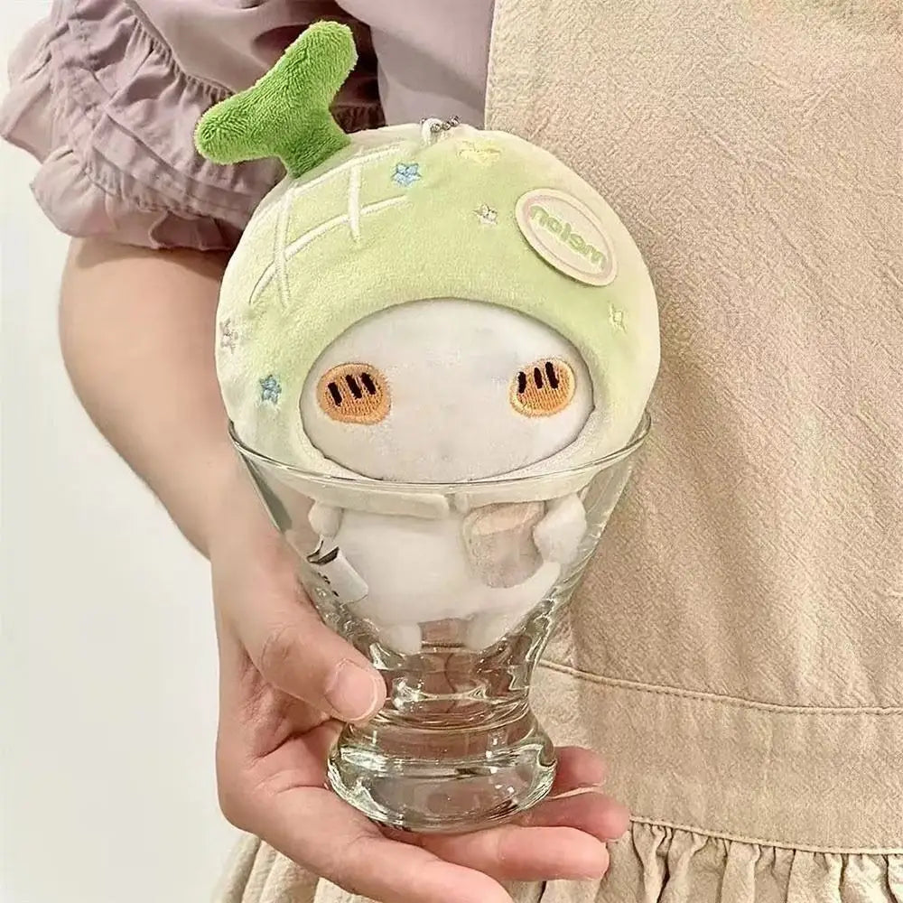 A soft light green plush doll headgear animal costume with embroidered orange eyes and a small green leafy top sits inside a clear glass cup measuring 10cm.