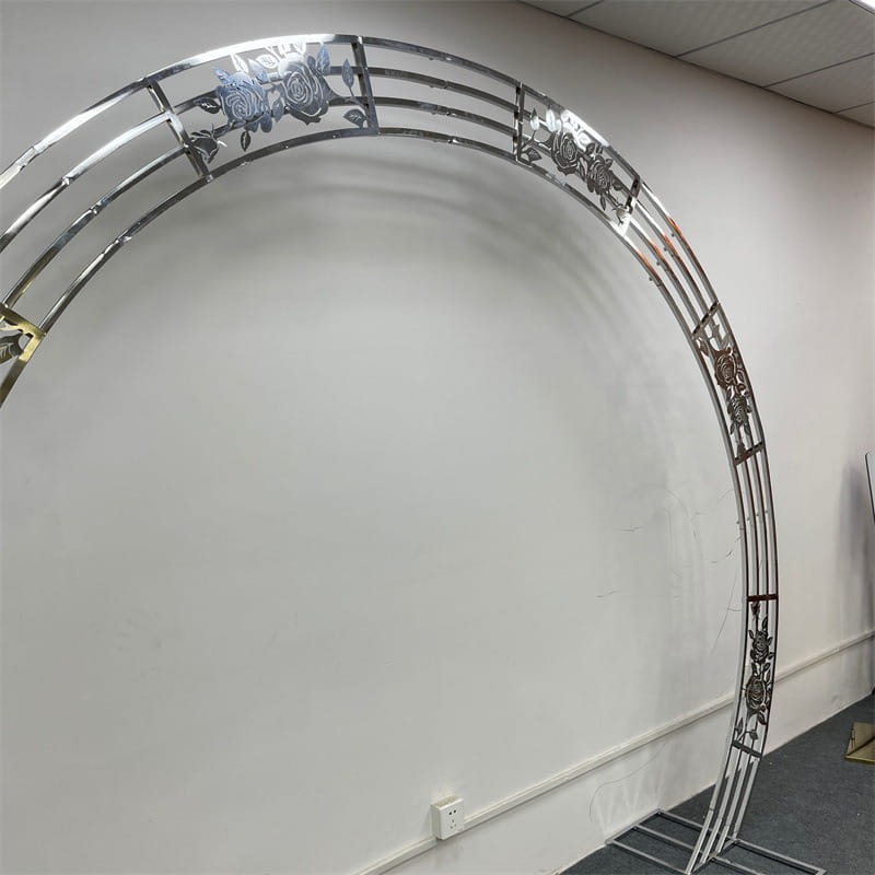 arche de mariage en métal chromé brillant avec des découpes de roses complexes le long de son cadre incurvé utilisée comme support de fond d’arche de mariage dorée pour des réceptions.