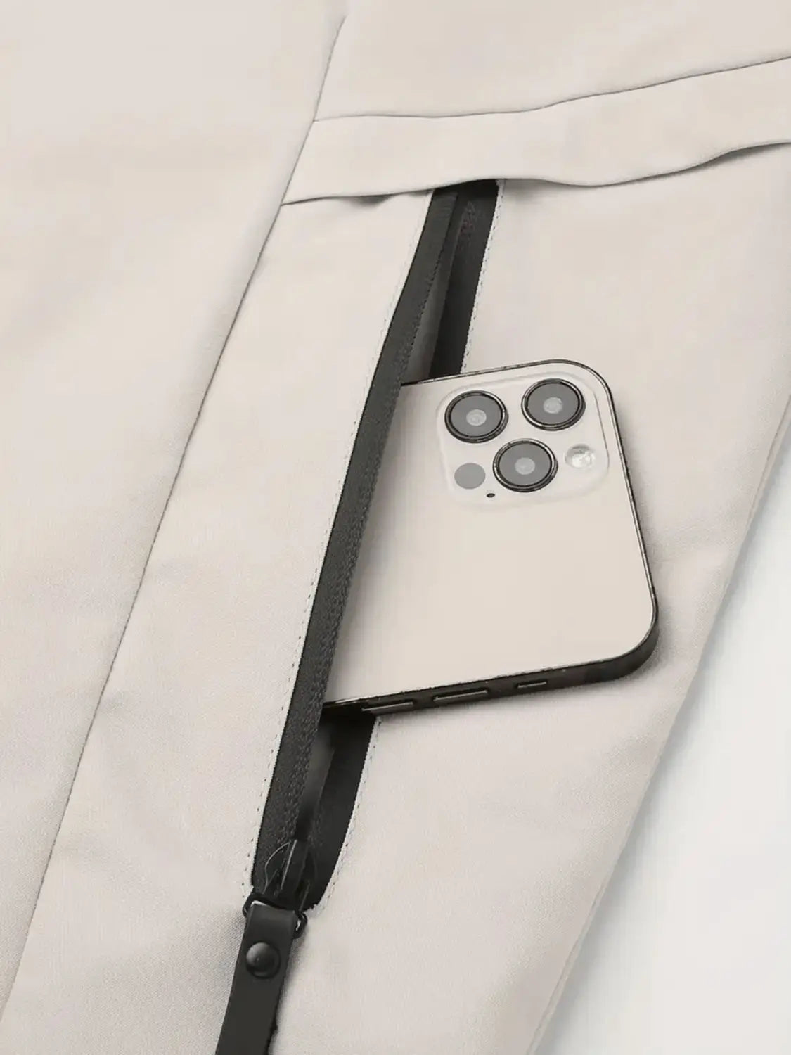 A sleek white iPhone with a triple-camera system rests partially inside the zipper of a light beige men off road hooded jacket windproof casual outerwear.