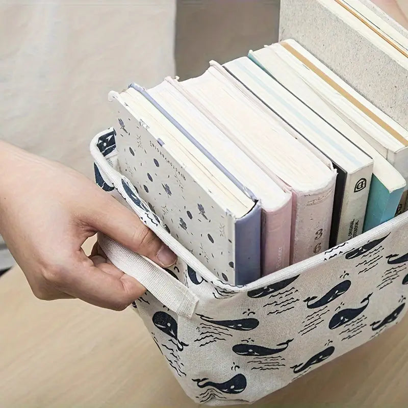 Panier de rangement pliable en tissu beige avec un motif de baleines noires et d’ondes, ainsi que des poignées blanches robustes pour un usage polyvalent en boîte de rangement.