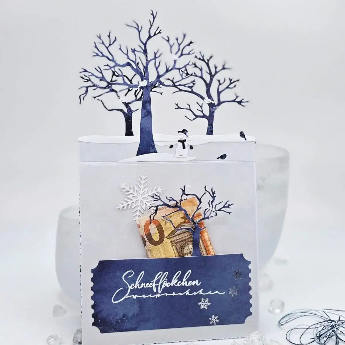 A white paper Christmas gift card features a winter scene with blue cutout trees a snowman and snowflakes created with winter animal metal cutting dies for scrapbooking.