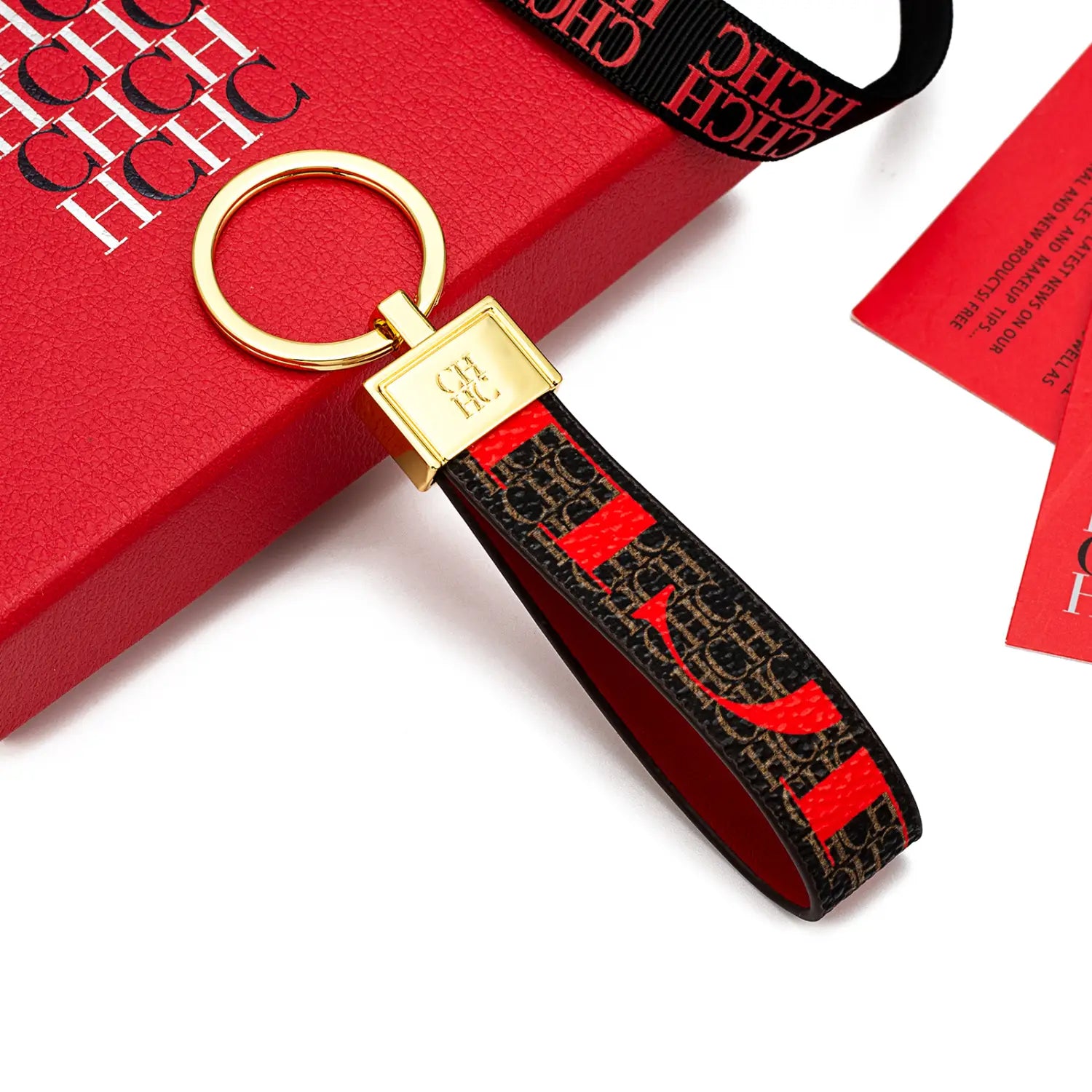 Gold-toned leather logo keychain with a black and red patterned fabric strap featuring a repeating hchc monogram design, ideal as a designer fashion accessory gift.