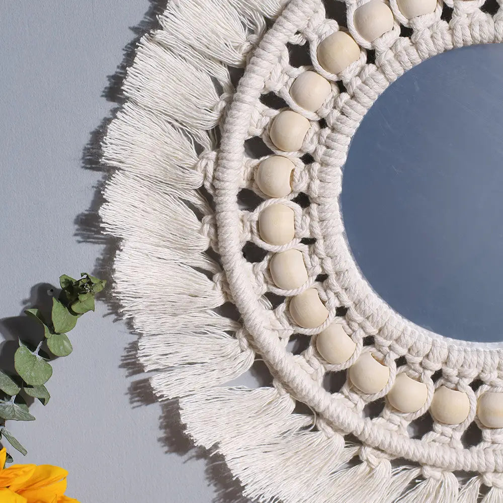 A cream-colored round macrame wall mirror with a decorative frame featuring wooden beads and fringe for boho style decorative mirrors.