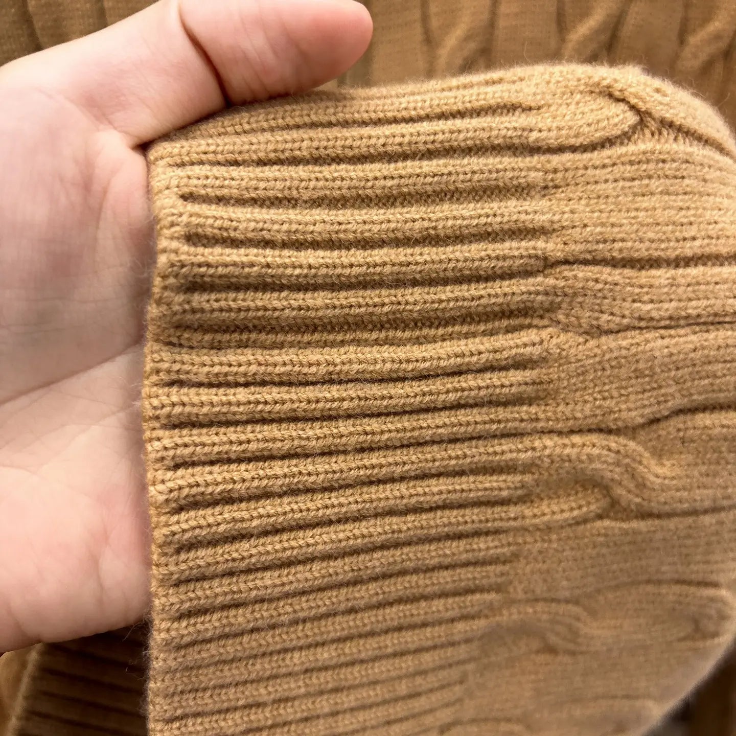 Nahaufnahme einer Hand, die das gerippte, camel-farbene Strickbündchen eines Herren-Designer-Sweaters aus Zopfmusterstrick mit halbem Reißverschluss hält.