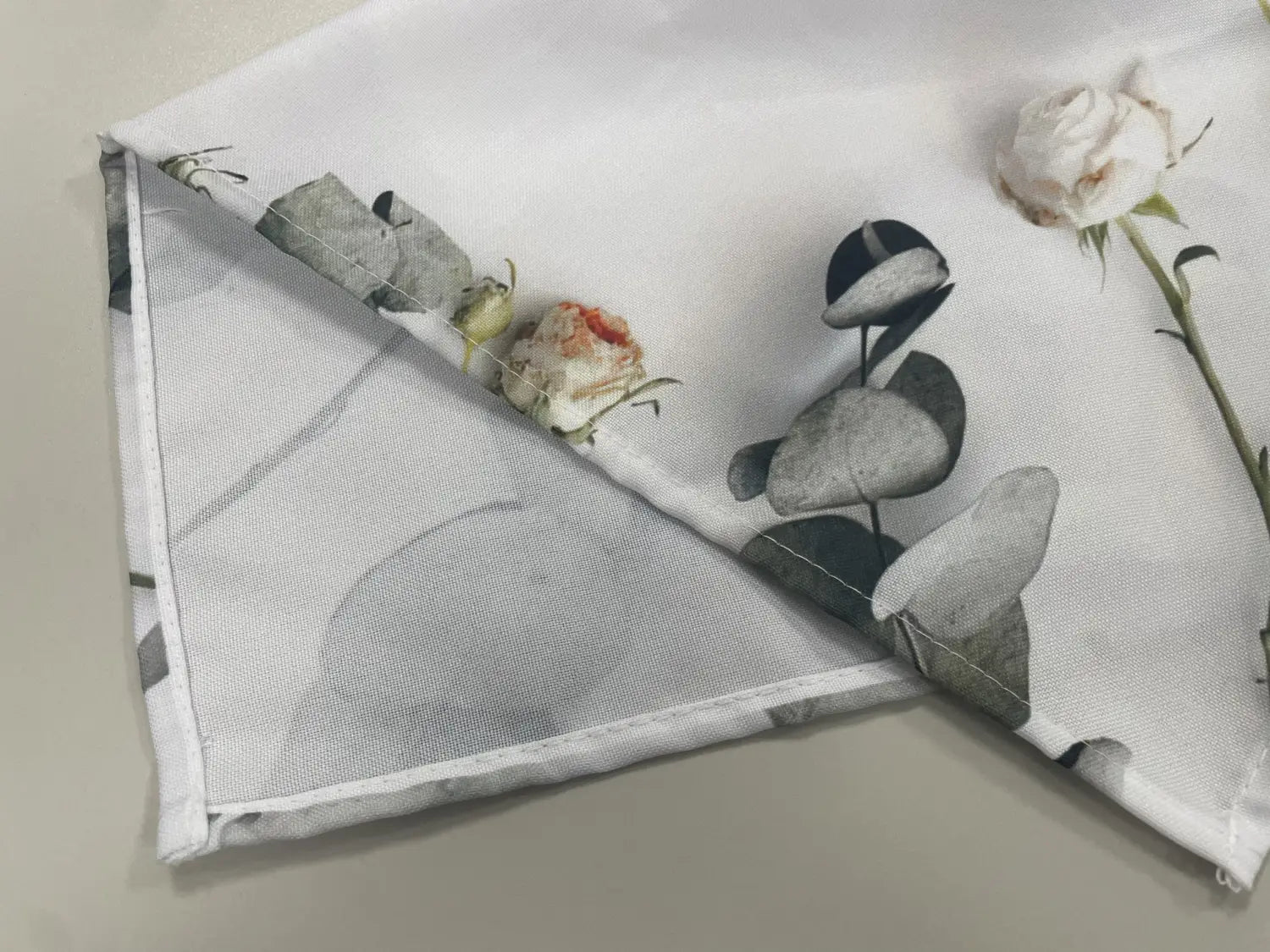 Weiße Stoff-Serviette mit zartem Blumenmuster, das gedämpfte graue Eukalyptusblätter und sanfte weiße Rosen zeigt, ergänzt den Vintage-Weihnachtsmann-Tischläufer im rustikalen Landhausstil.