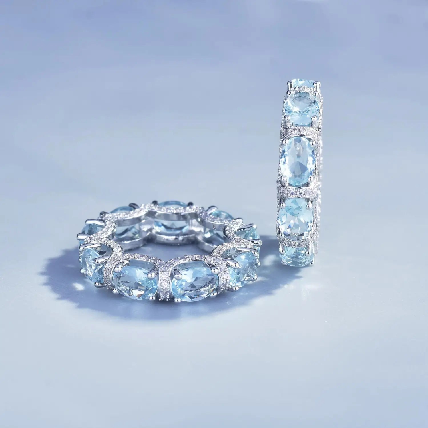 A pair of elegant silver-toned aquamarine crystal bridal rings featuring oval-cut sky blue gemstones set in a pave design with one ring displayed horizontally and the other vertically.