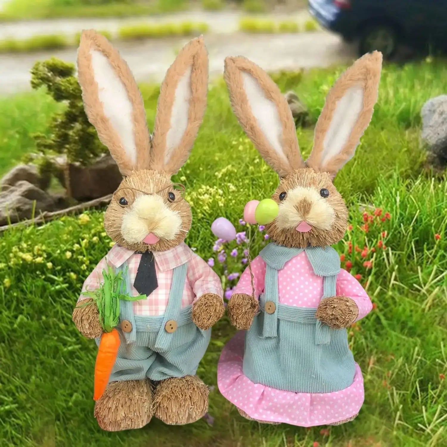 Two charming 35cm straw Easter rabbit figurines featuring one dressed in a pink plaid shirt with blue overalls holding a carrot and the other in a pink polka-dot dress with matching overalls, both with long tan ears with white interiors.