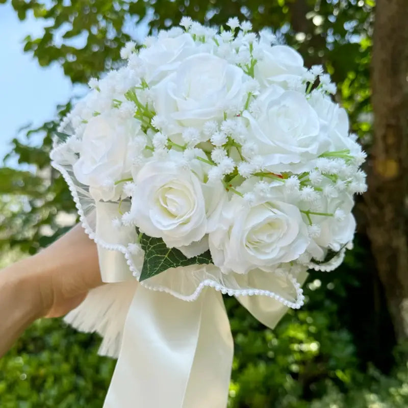 A delicate white silk rose wedding bouquet adorned with baby’s breath and tied with a satin ribbon featuring lace and pearls.