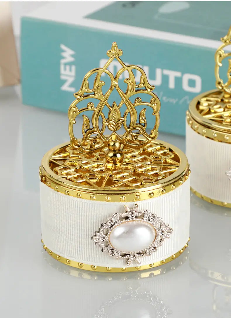 Boîte à bonbons de mariage décorative blanche cannelée avec bordure dorée et couvercle en filigrane doré orné, featuring une perle centrale, entourée d’éléments rehaussés à la finition ton argent, utilisée comme boîte à bonbons de mariage dorée décorative, contenant une perle-bonbon en guise de cadeau.