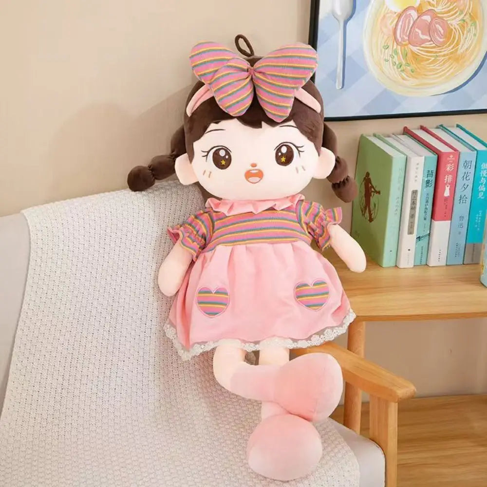 Plush doll 45cm cute stuffed animal toy with brown braided pigtails wearing a pink dress with rainbow-striped heart patches and matching striped sleeves and a large rainbow-striped bow on her head.