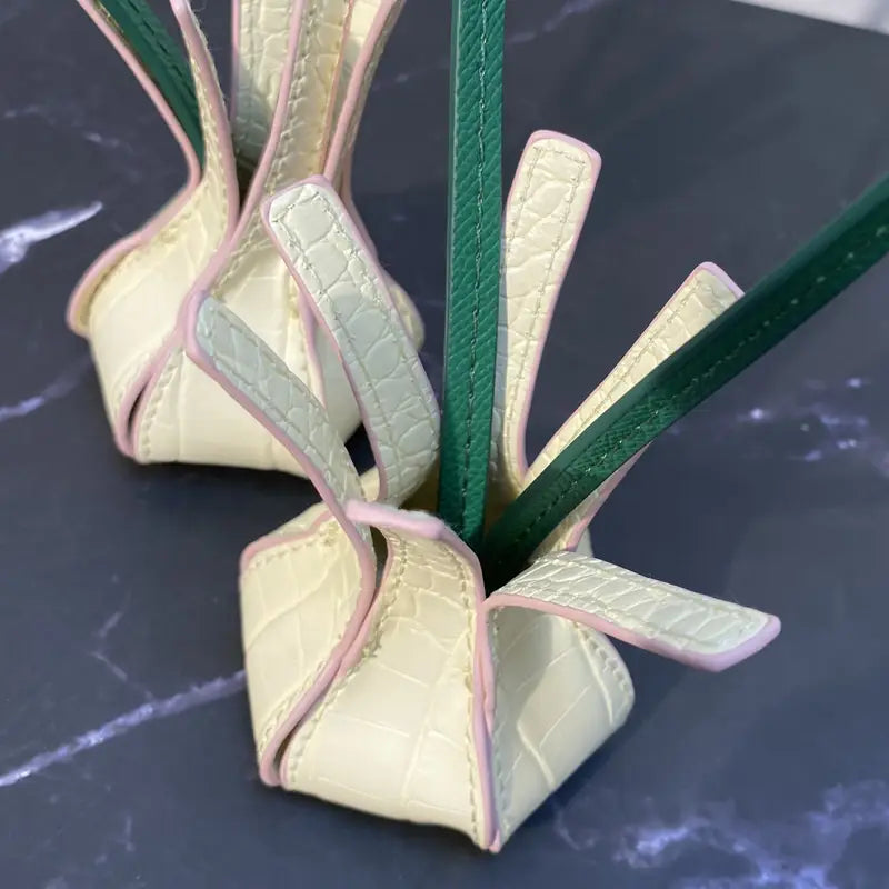 Un charm pour sac à main en cuir végétal, couleur jaune pastel, gaufré façon crocodile, avec une couture rose et des lanières vertes formant une poignée d’ornement de sac à main au motif fleuri.