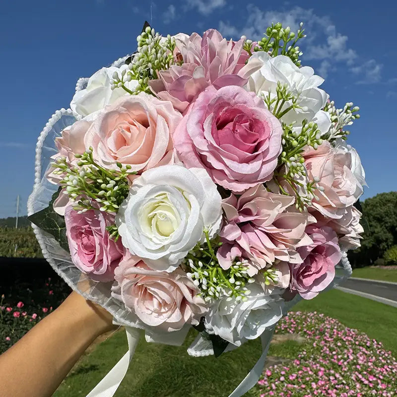 A delicate bouquet of white silk artificial roses with soft pink and white hues accented with greenery and wrapped in a sheer lace ribbon suitable for weddings.