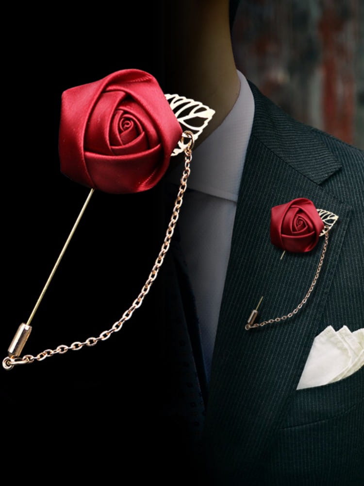 Boutonnière élégante de rose bordeaux composée d’un ruban rouge en satin, rehaussée d’une chaîne à tons dorés et d’un accent feuillu, conçue comme une épingle de revers.