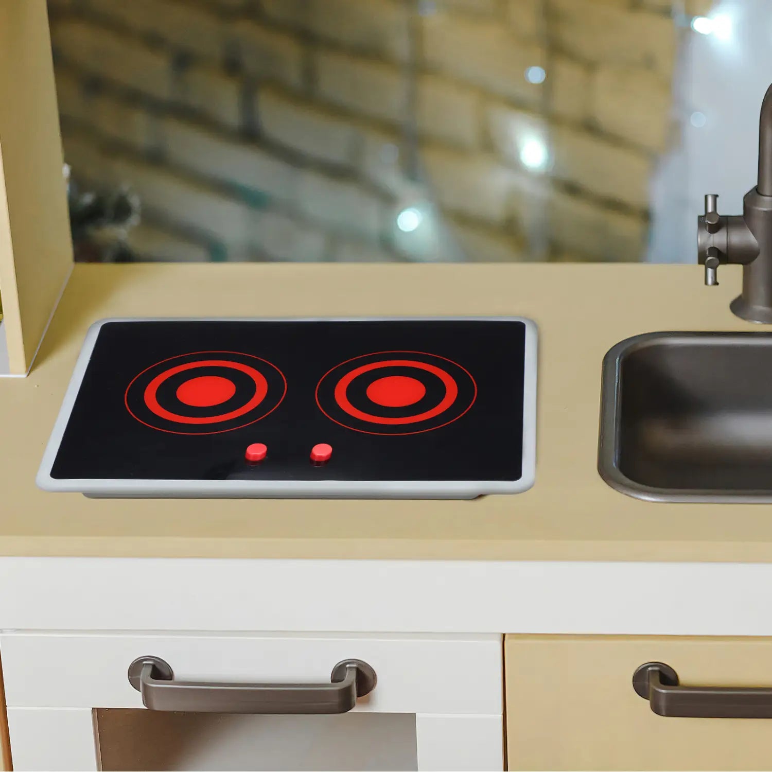 Schwarzer Spielzeug-Induktionsherd mit roten kreisförmigen Heizelementen und zwei kleinen roten Bedientasten, eingelassen in eine beige Arbeitsplatte neben einem Edelstahlspülbecken, mit Sound- und Lichtfunktionen.