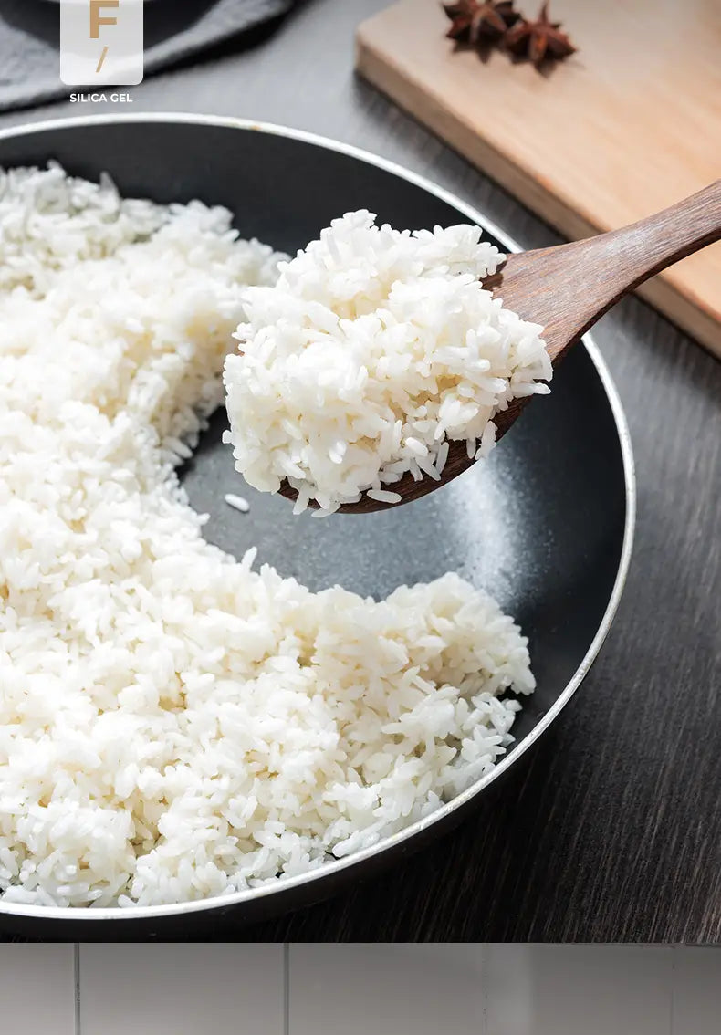 A wooden spoon from a wooden kitchen utensil set lifts a scoop of fluffy white cooked rice from a black non-stick cookware frying pan.