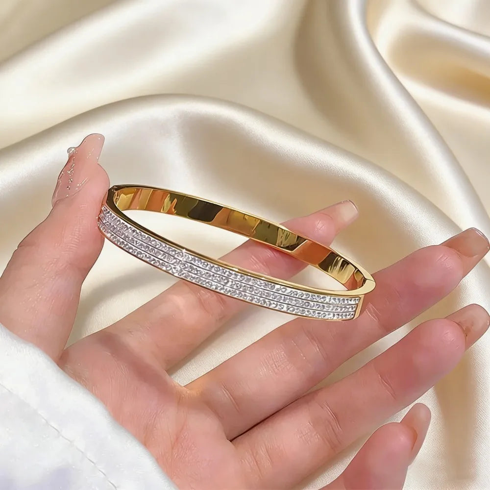 Gold-toned stainless steel bangle bracelet for women featuring three parallel rows of sparkling clear crystals and Roman numeral design.