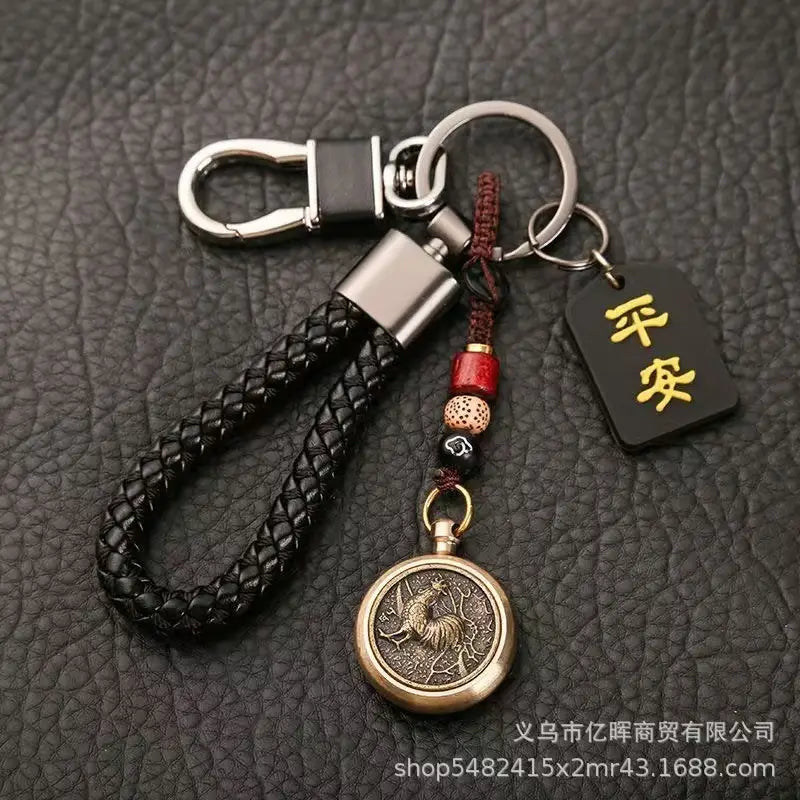 赤と黒のビーズでアクセントを施し、刻印された「鶏」のデザインをあしらった真鍮色の円形ペンダントを備えた黒の編みレザー製キーチェーン。金色の漢字で記された小さな黒いタグ付き（中国の干支をモチーフにした編みレザー製ラッキーペンダント）。