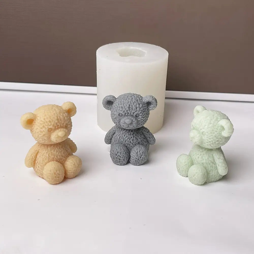 drei kleine Mini-Teddybär-Kerzenfiguren in beige-grau und hellgrün neben einer weißen zylindrischen Silikonform zur Herstellung von Kerzen, von vorne aus gesehen