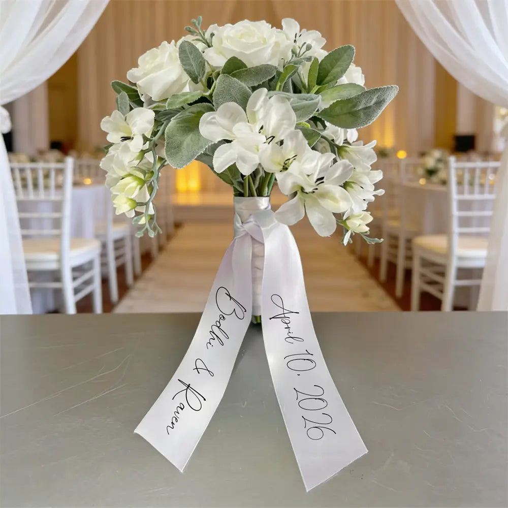Un délicat bouquet de mariée artificiel blanc, aux pétales au toucher doux évoquant la soie et aux feuilles vert sauge, noué avec un ruban de satin personnalisé portant les noms propres et la date, pour un usage lors du mariage