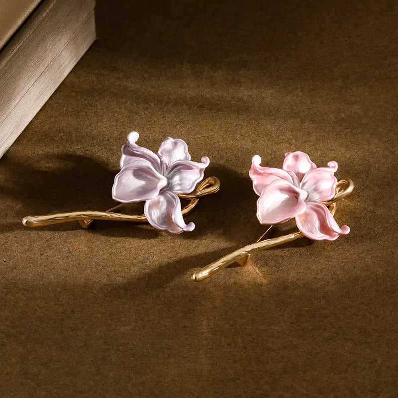Une paire de broches-épingle émaillées en forme de fleur de lotus, présentant des fleurs en porcelaine peintes à la main dans des teintes lavande et rose douces, montées sur des tiges torsadées à ton doré