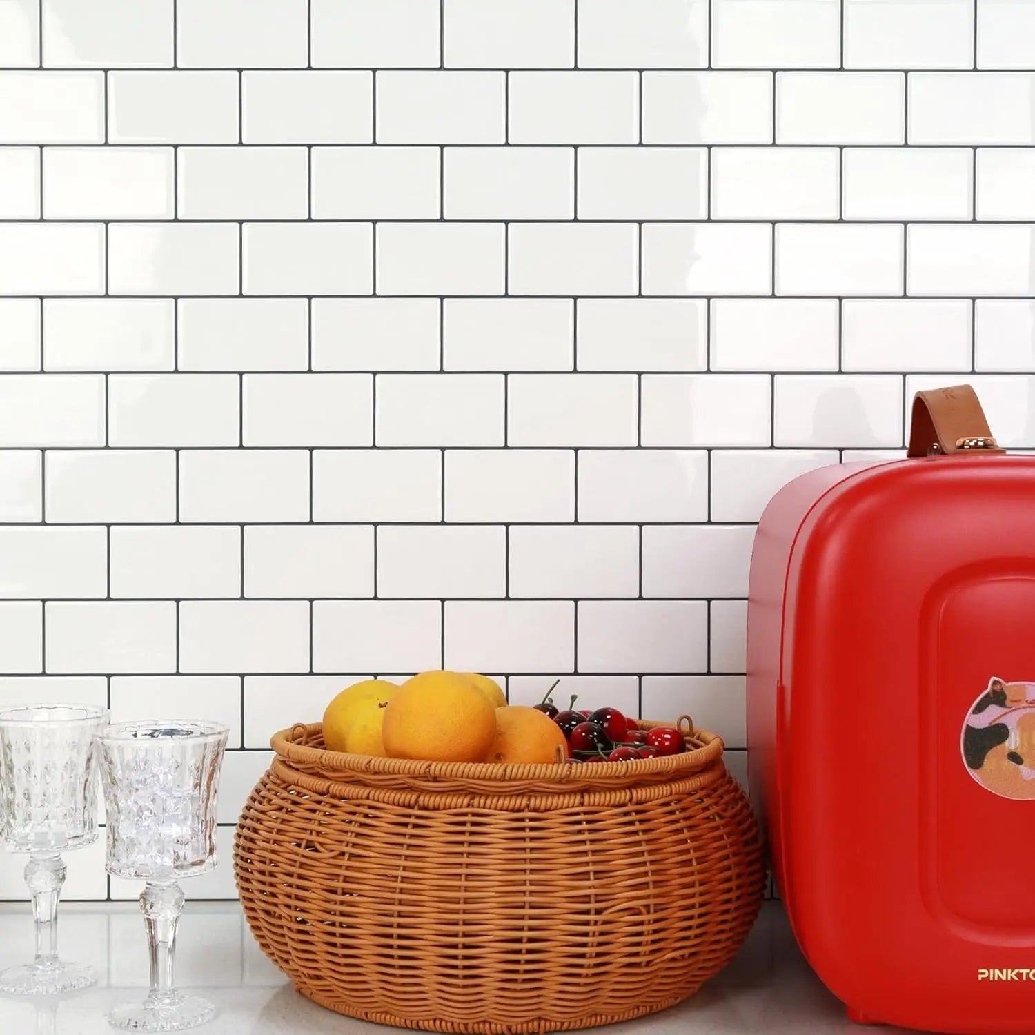 a woven wicker basket with a natural brown hue and intricate interlaced pattern filled with oranges and cherries placed against a peel and stick backsplash tile glossy white subway highlighting direct contact with fresh paint surface.