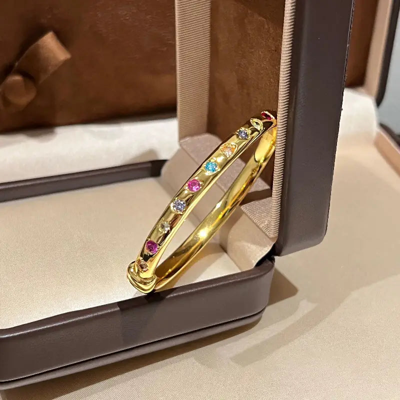 Ein Armreifarmband aus goldfarbenem Multigemsteinschmuck, das eine lebendige Vielfalt an farbigen Edelsteinen elegant in einer offenen Schmuckschatulle zur Schau stellt.