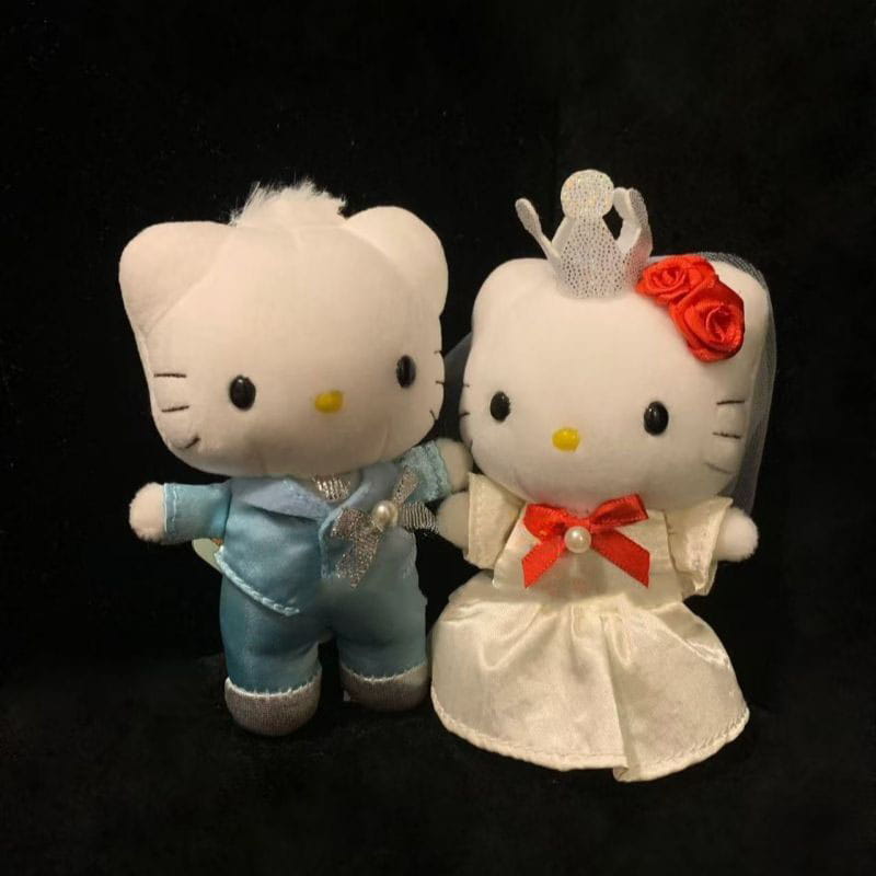 porte-clés en peluche couple de mariage Hello Kitty, représentant des figurines en peluche blanches habillées d’un costume en satin bleu clair avec un passepoil argenté, et d’une robe en satin ivoire décorée de roses rouges, d’un nœud en perles et d’un voile délicat.