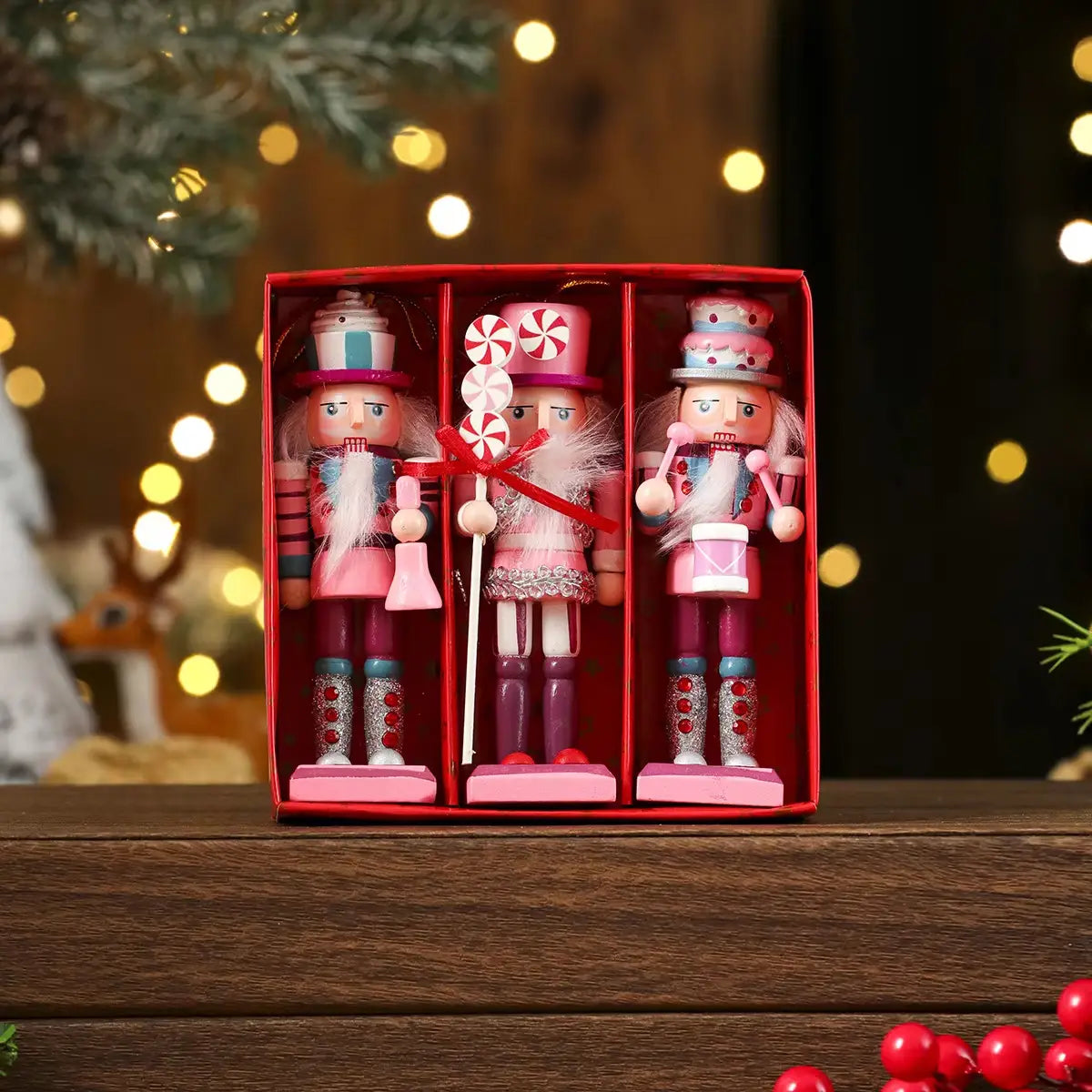 Tres figurines de cascanueces de madera en tonos rosa y morado con accesorios temáticos de caramelos, exhibidos en una caja roja como decoración de árbol de Navidad.