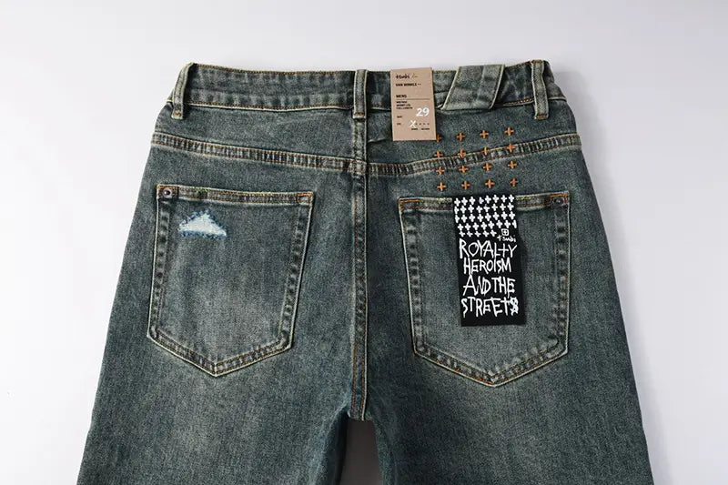 men’s distressed blue acid wash denim shorts shown from the back side featuring a distressed patch on the left back pocket and a black graphic patch with white text reading royalty heroism and the streets on the right pocket.