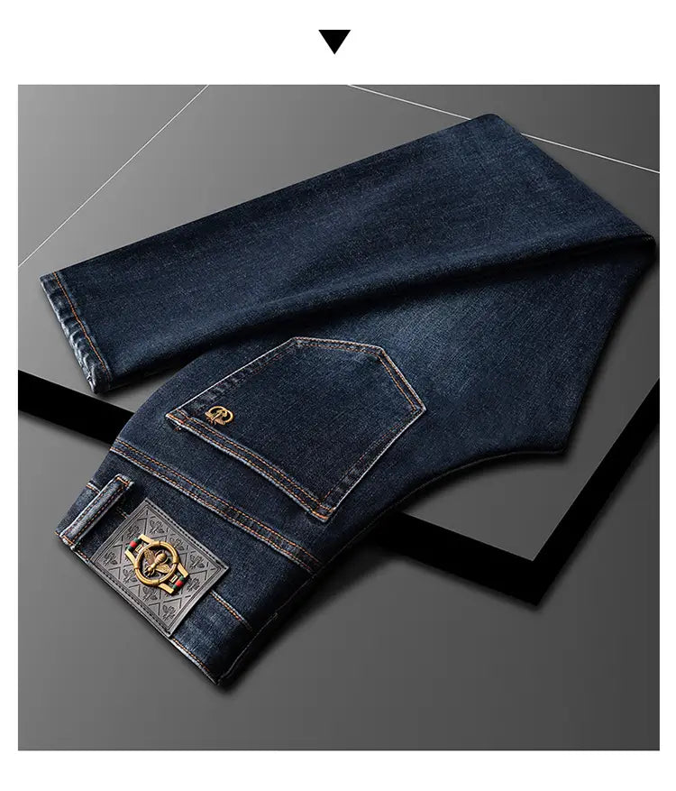 Slim fit dark blue denim jeans for men shown from the back with a decorative gold-toned logo patch on the pocket and an ornate metallic brand plaque at the waistband.