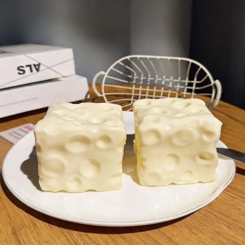 Two creamy off-white cube-shaped cheese block stress relief squeeze toys with a textured surface mimicking swiss cheese holes sit on a white plate.