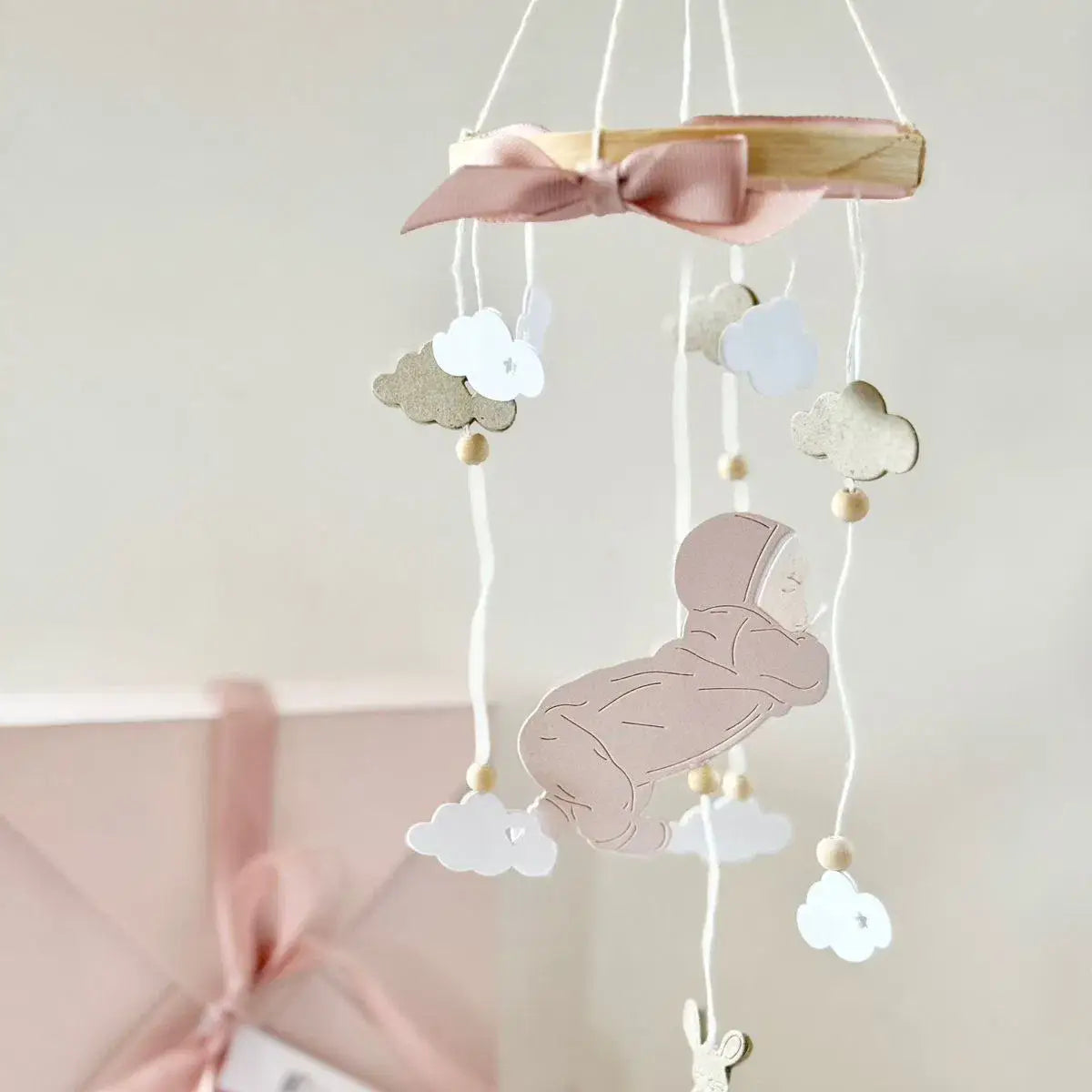 Oster-Ei-Hasen-Babyschneidvorlagen mit einem zarten rosa Band, das an einem hölzernen Mobile mit ausgeschnittenen Wolken gebunden ist, sowie einer schlafenden Babykontur, die an weißen Schnüren hängt.