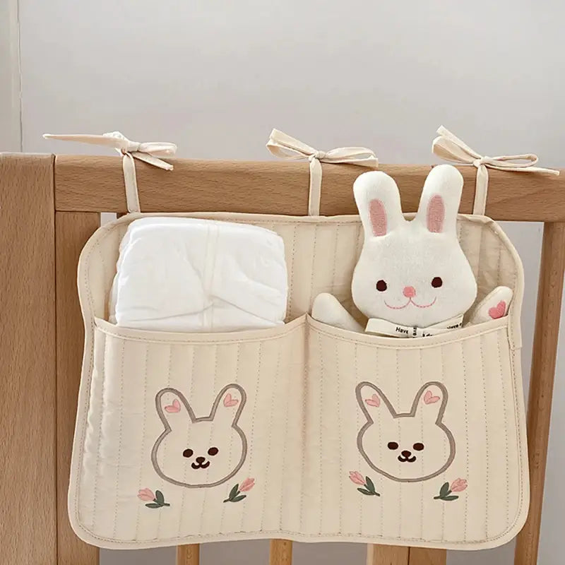 クマの刺繍が施されたクリーム色のキルティング製ベビーカー用オーガナイザー袋で、刺繍のうさぎの顔とピンクの花柄のディテールがあり、ポケットが2つでオムツとふわふわの白いうさぎのおもちゃを収納できます。