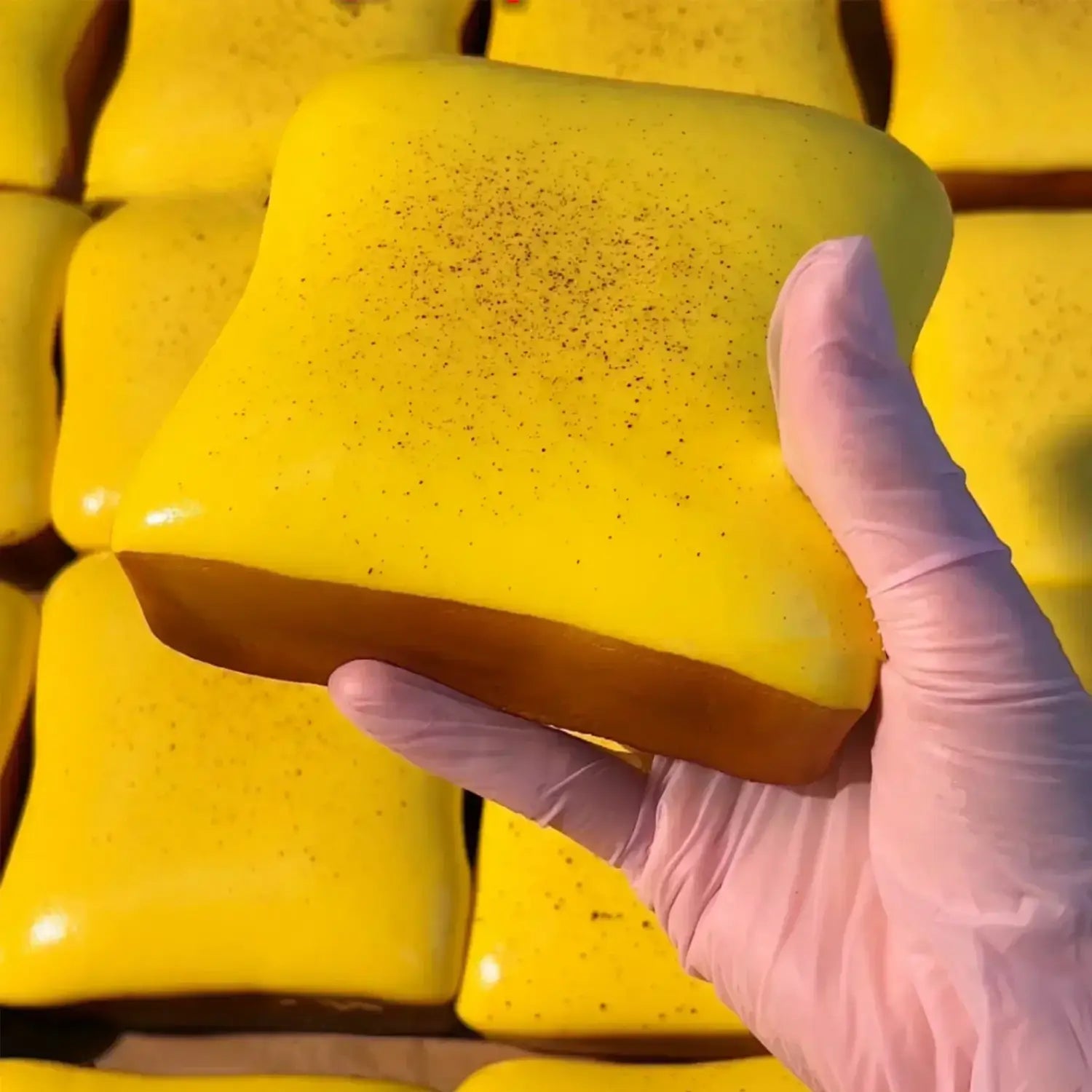 Une main portant un gant rose maintient un jouet à mâcher et à malaxer en forme de fromage jaune vif, brillant, conçu pour soulager le stress, texture souple et molle, avec une base brune et un dessus tacheté, conçu pour apaiser le stress lié au jouet.