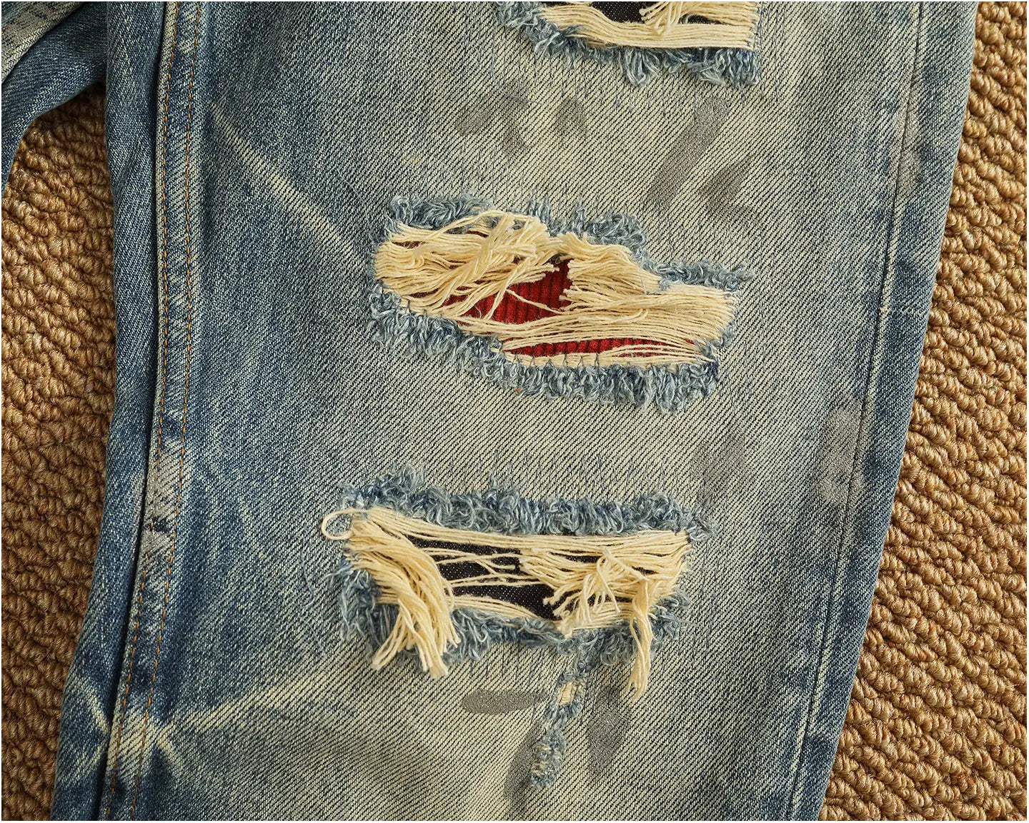 Distressed patchwork denim jeans men featuring blue denim fabric with frayed ripped holes revealing red and black inner fabric.