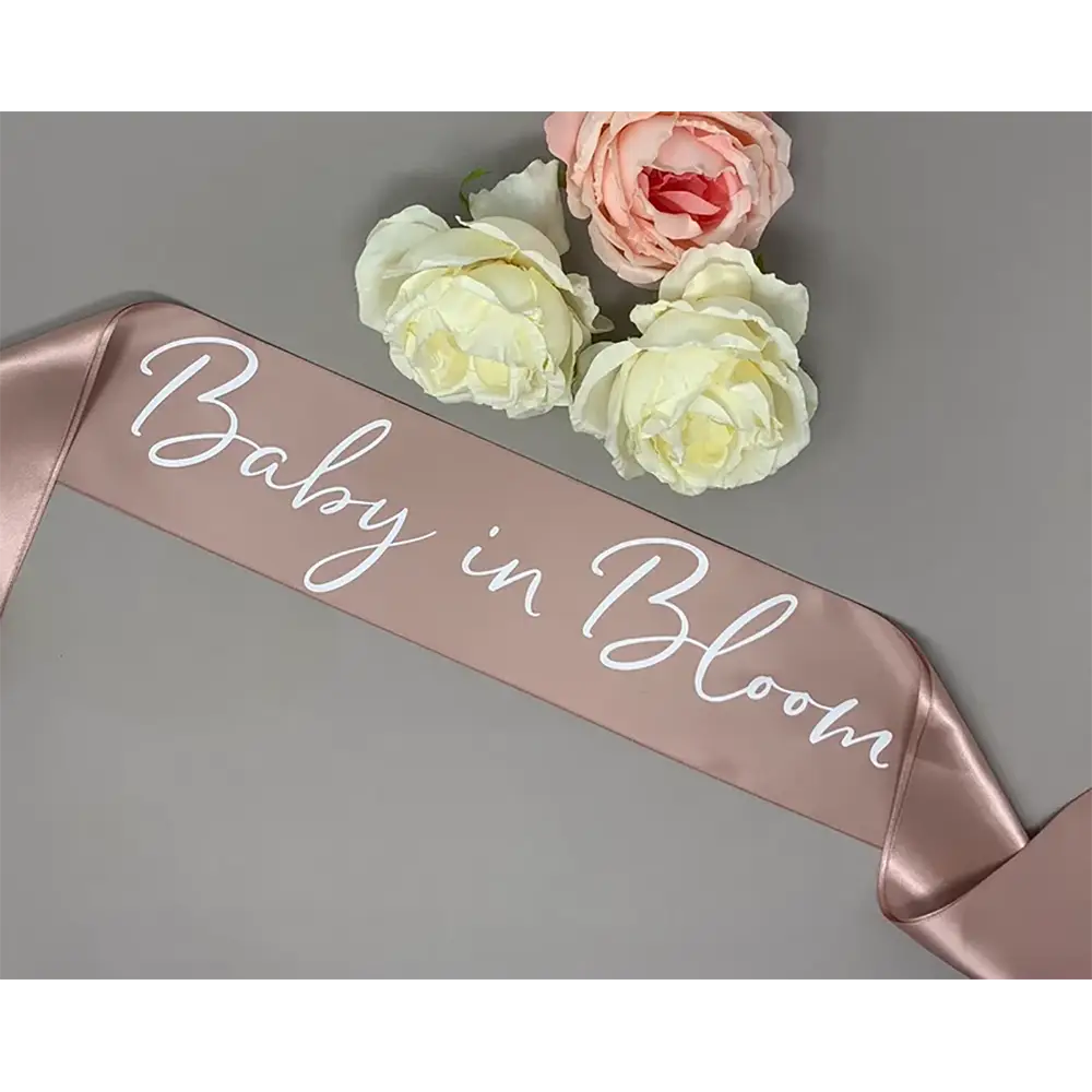Une écharpe de baby shower en satin rose doré pâle avec « baby in bloom » écrit en script cursif blanc pour la future maman, modèle en satin blush rose pâle.