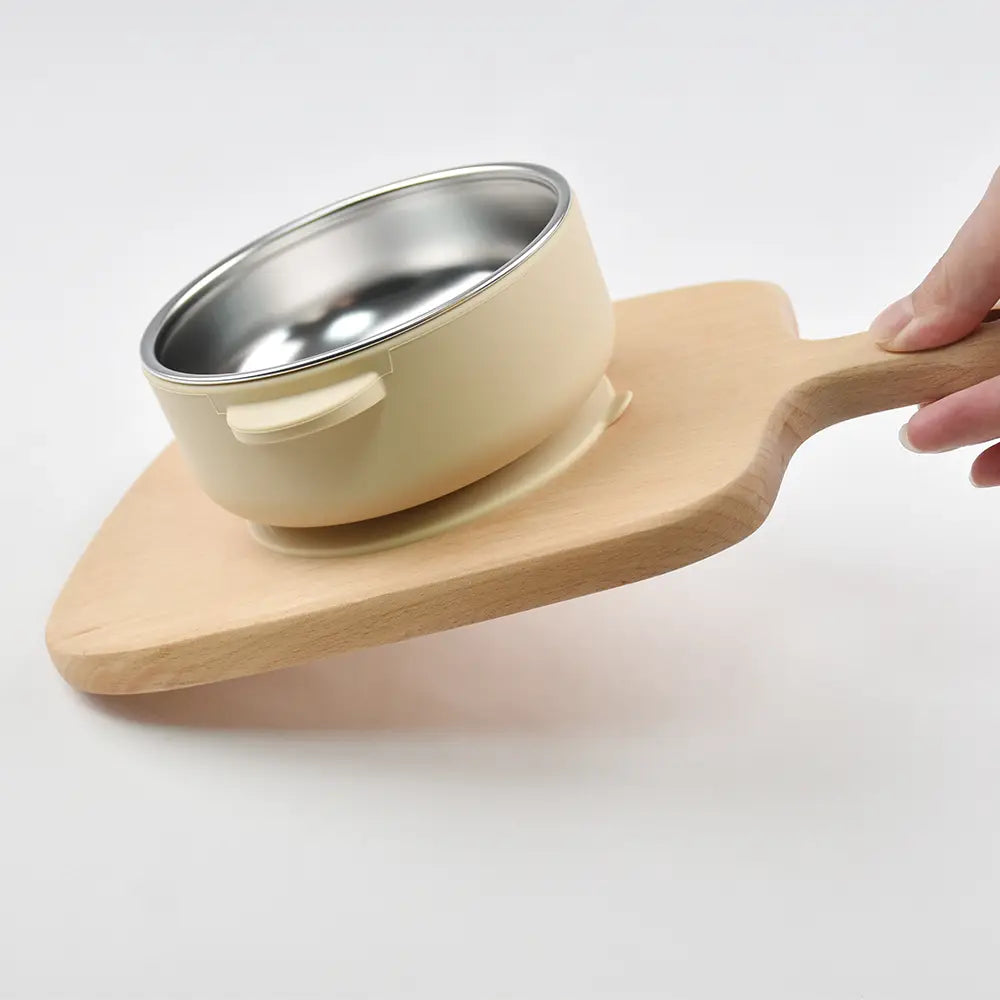 Cremefarbene Baby-Trainingsschüssel mit Edelstahlinnenbereich und kleinem Griff, abgelegt auf einem Holz-Servierbrett; konzipiert für das Zufüttern und Füttern.