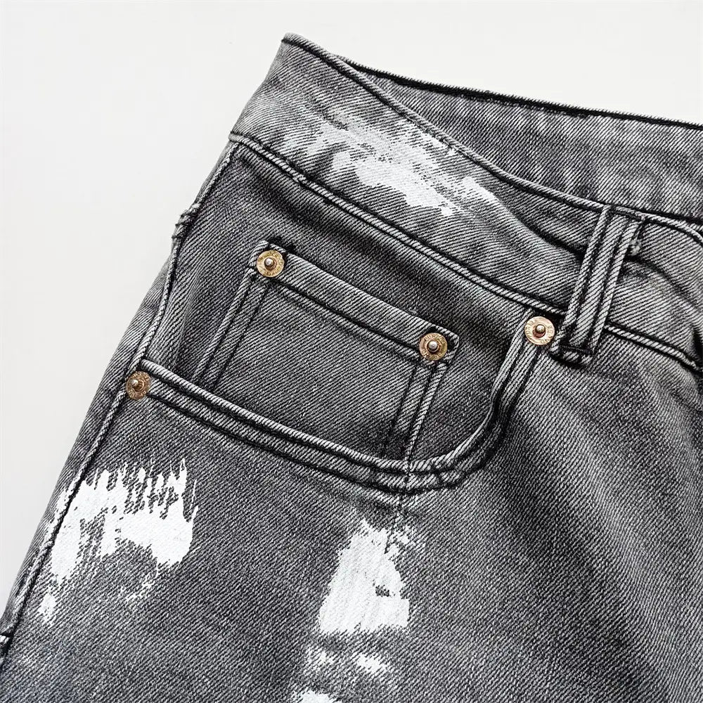 Jeans skinny déchirés pour hommes en denim noir déchiré, avec des accents d’éclaboussures de peinture blanches et des fermetures par boutons ton or.