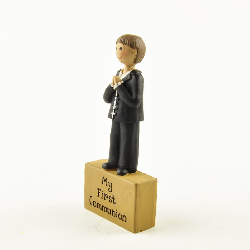une figurine en résine d’un garçon en tenue noire tenant des perles de chapelet se tient sur une base en bois gravée « ma première communion », conçue comme décoration de table pour la première communion.