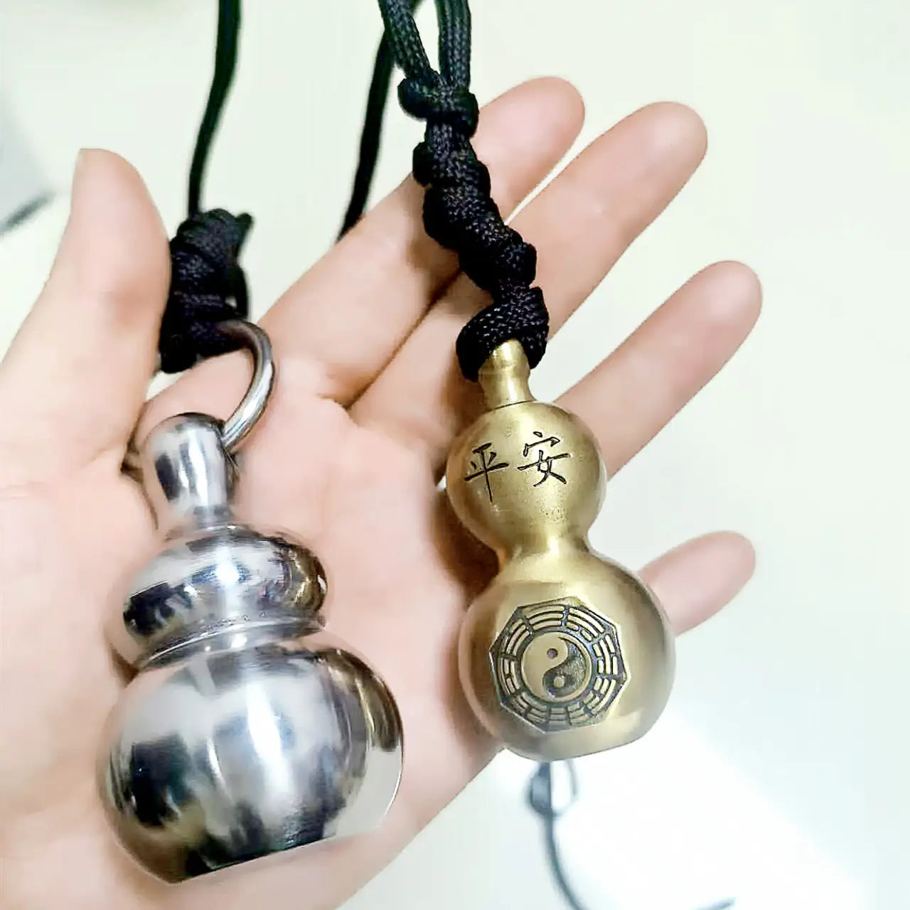 Two metallic brass gourd self defense pendant necklaces in silver and gold tones featuring engraved Chinese characters peace and a yin-yang symbol respectively, each attached to black braided cords.