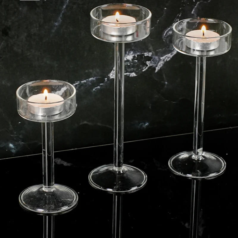 Tres portavelas de pilar de vidrio transparente, con tallos esbeltos y bases circulares, cada uno con una vela tipo té encendida, para una decoración moderna de mesa.