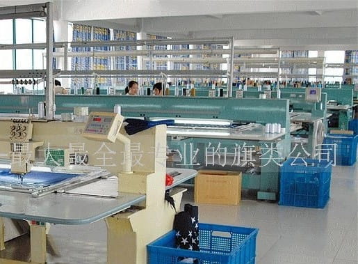 Reihen von türkisfarbenen industriellen Nähmaschinen mit digitalen Anzeigen, die auf dem Fabrikboden neben blauen Kunststoffkisten angeordnet sind und die Produktion von Deutschland-Flaggenprodukten mit Stangenhülse unterstützen.