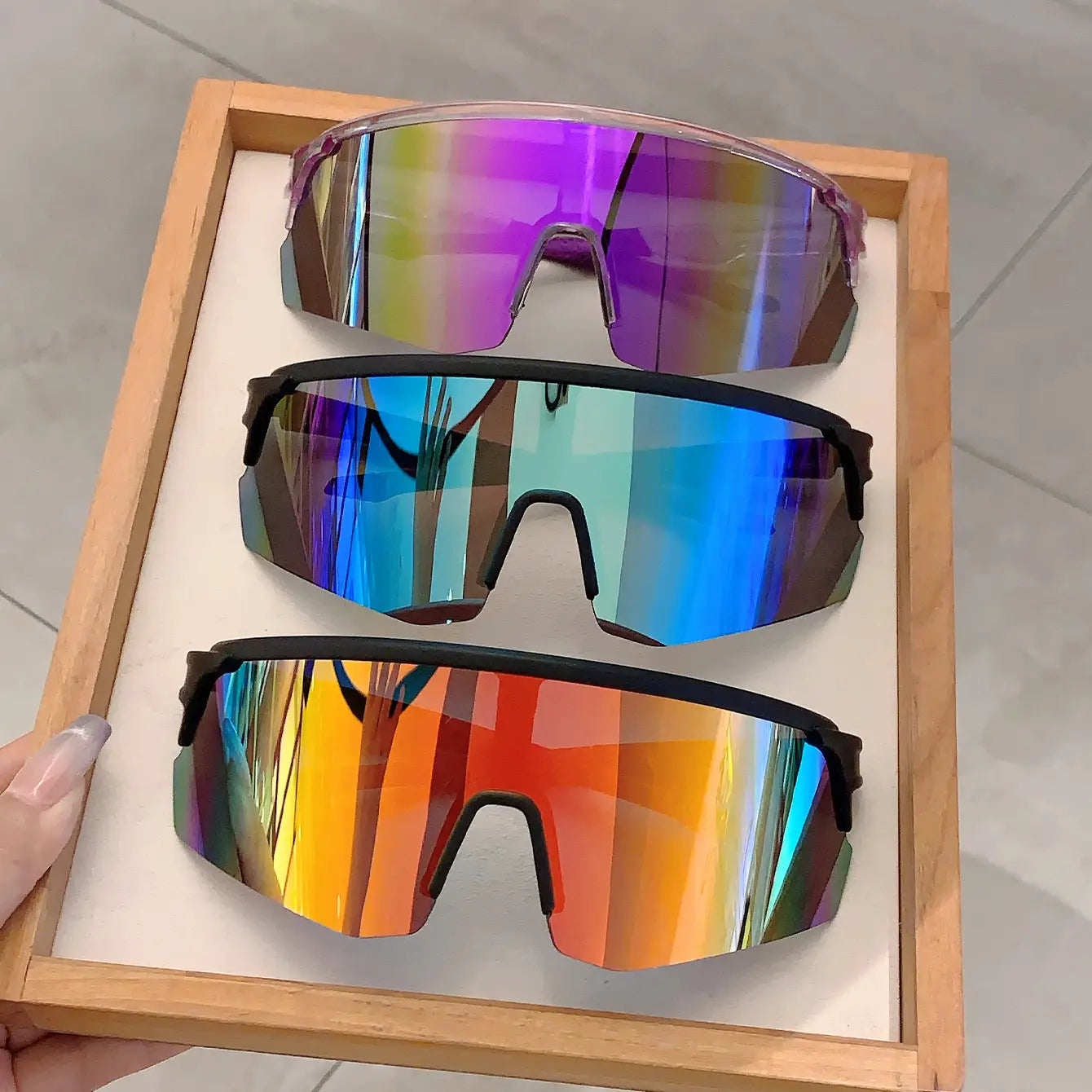 Trois paires de lunettes de soleil de sport surdimensionnées, avec des verres enveloppants irisés dégradés en tons violet, bleu et rouge orangé, encadrées par des branches noires élégantes et du plastique translucide, conçues comme des lunettes de cyclisme à effet miroir.