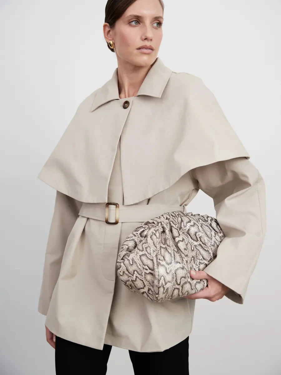 Abrigo tipo gabardina marrón claro de mujer ajustado con cinturón y superposición de capa, con una cartera con textura en color crema y estampado de serpiente negro, de diseño abullonado recogido