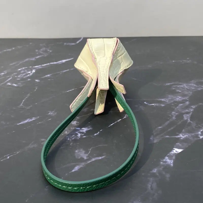 Une pochette délicate en cuir gaufré façon crocodile, couleur crème, avec une lanière vert vif et un design de nœud élégant, mise en scène comme charm pour sac à main en cuir végétal, ornement de sac à main de créateur.